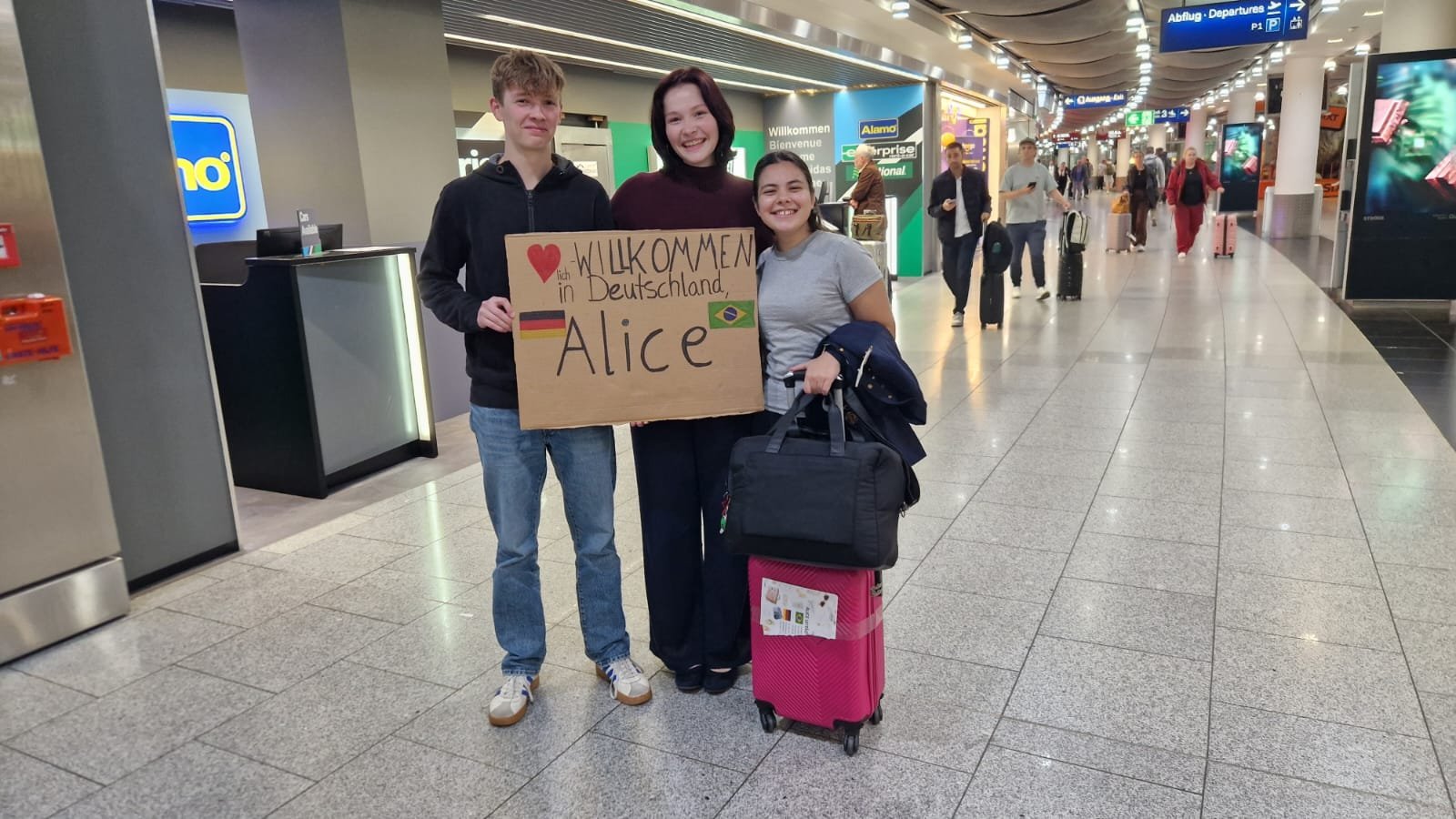 Alice na chegada à Alemanha, recebida por dois jovens com um cartaz de boas-vindas escrito Willkommen in Deutschland, Alice